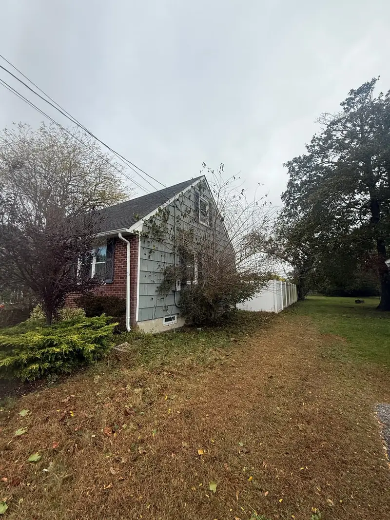 Side of a shingled Cape with overgrown shrubs on an overcast day, Patchogue, NY